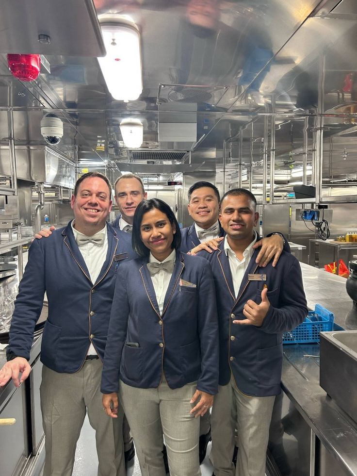 A group of five cruise ship crew members in matching uniforms smiling and posing together inside a stainless-steel ship kitchen or galley.